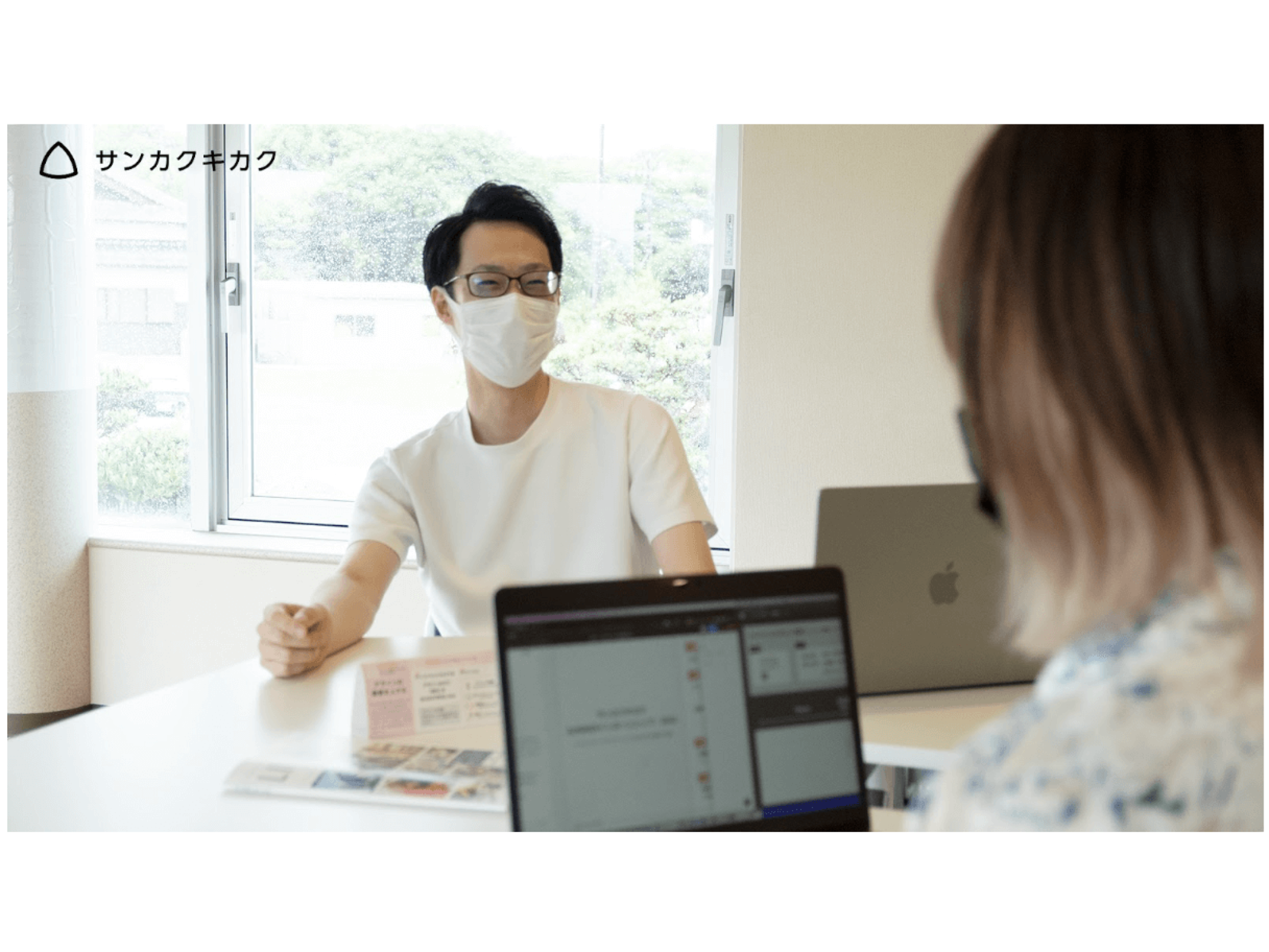 窓辺の机で、ノートパソコンを挟んで向かい合って話すマスク姿の男性と女性