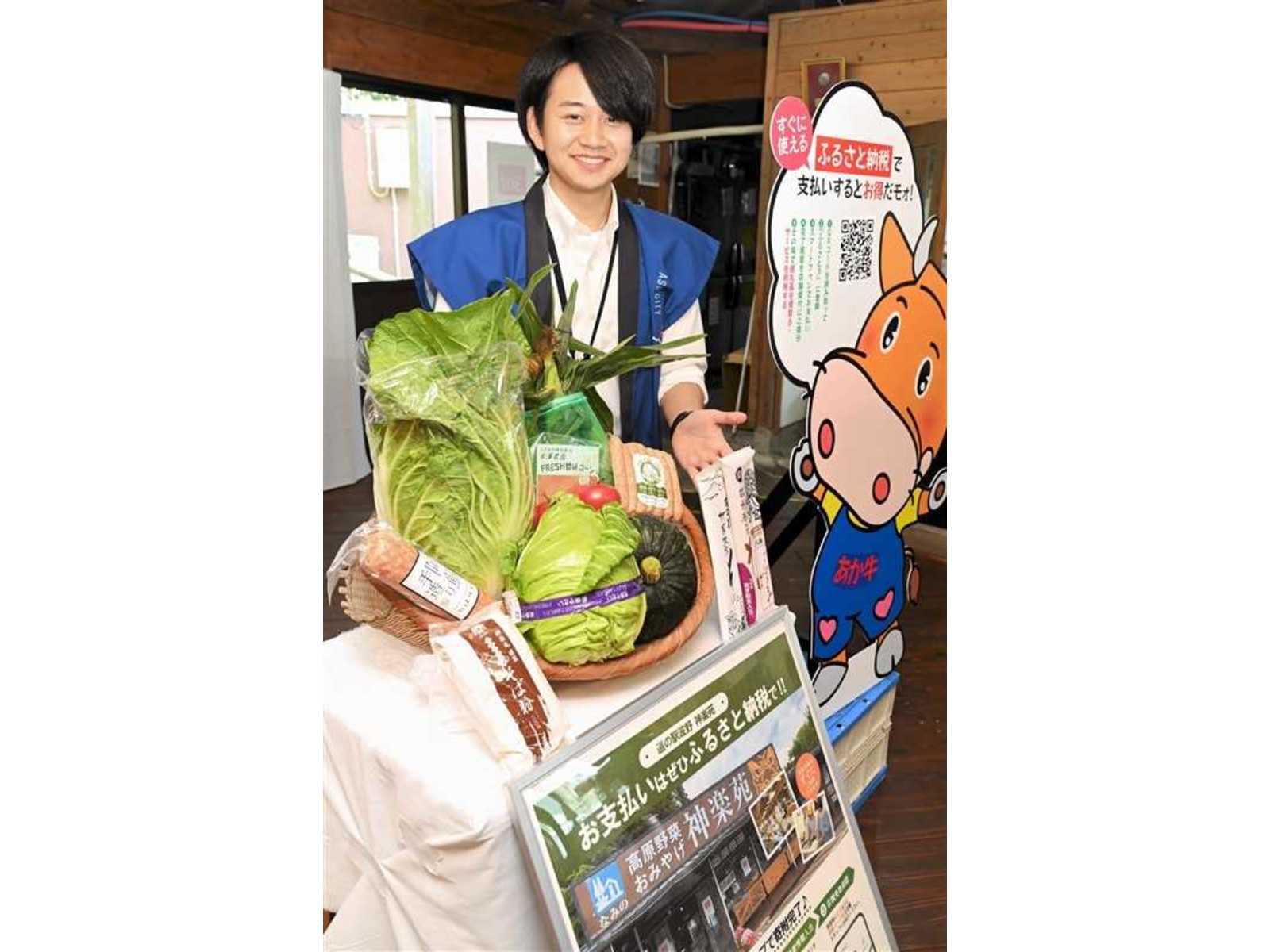 キャベツやかぼちゃを盛ったかごと「ふるさと納税」の案内パネルの横で、笑顔を見せる青いベストの男性