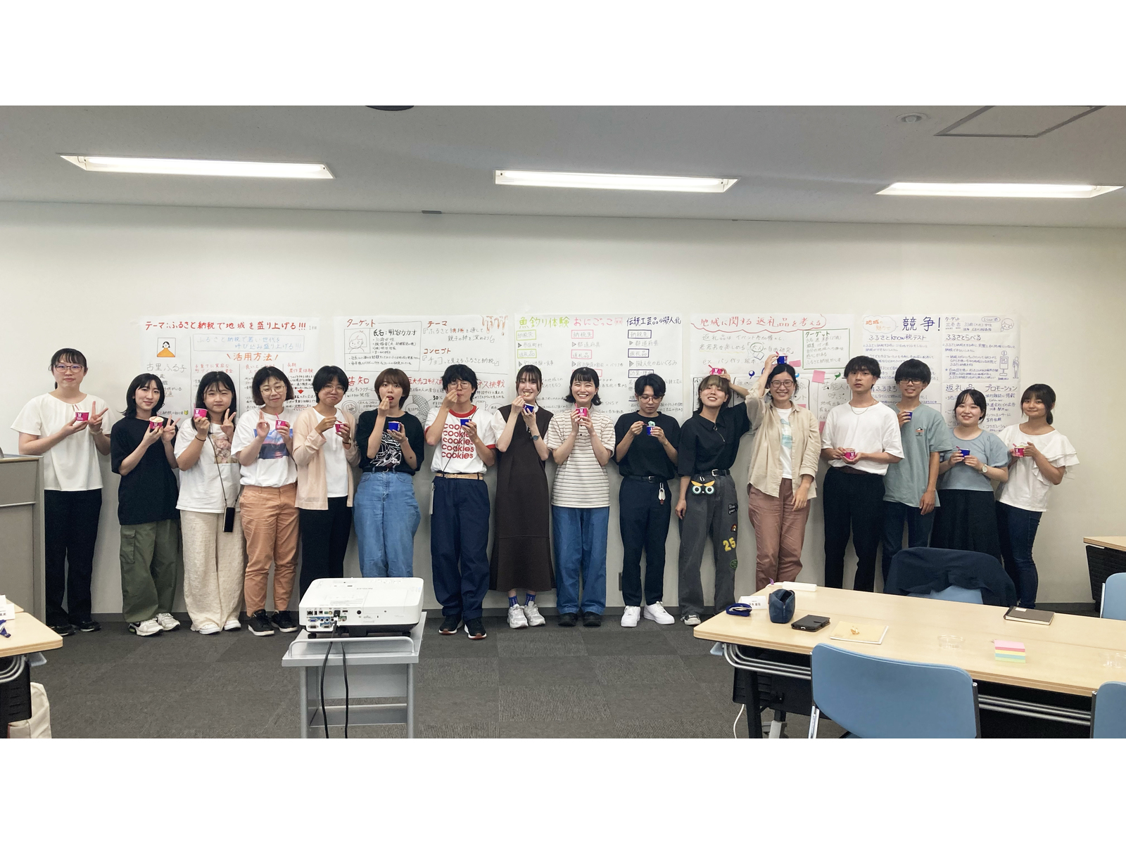 手書きの模造紙を貼った壁の前で、カップを手に並ぶ学生たち