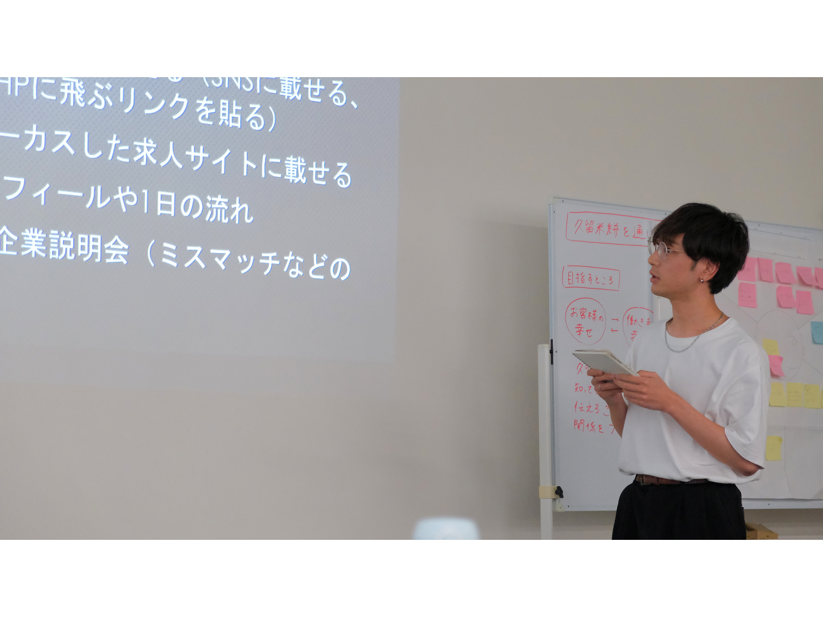 「求人サイトに載せる」「企業説明会」などを映したスクリーンの横で、タブレットを手に発表する学生