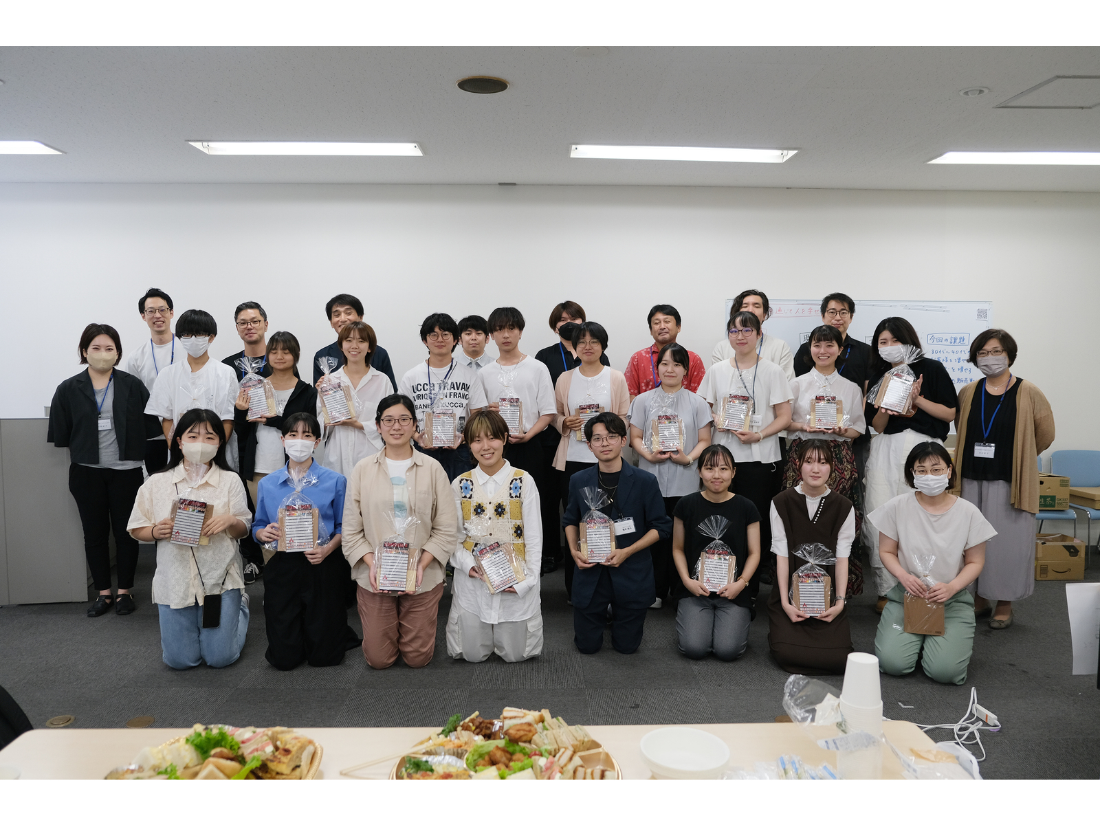 料理の並ぶ机の向こうで、包装された記念品を手に並ぶ学生たちと社員たち
