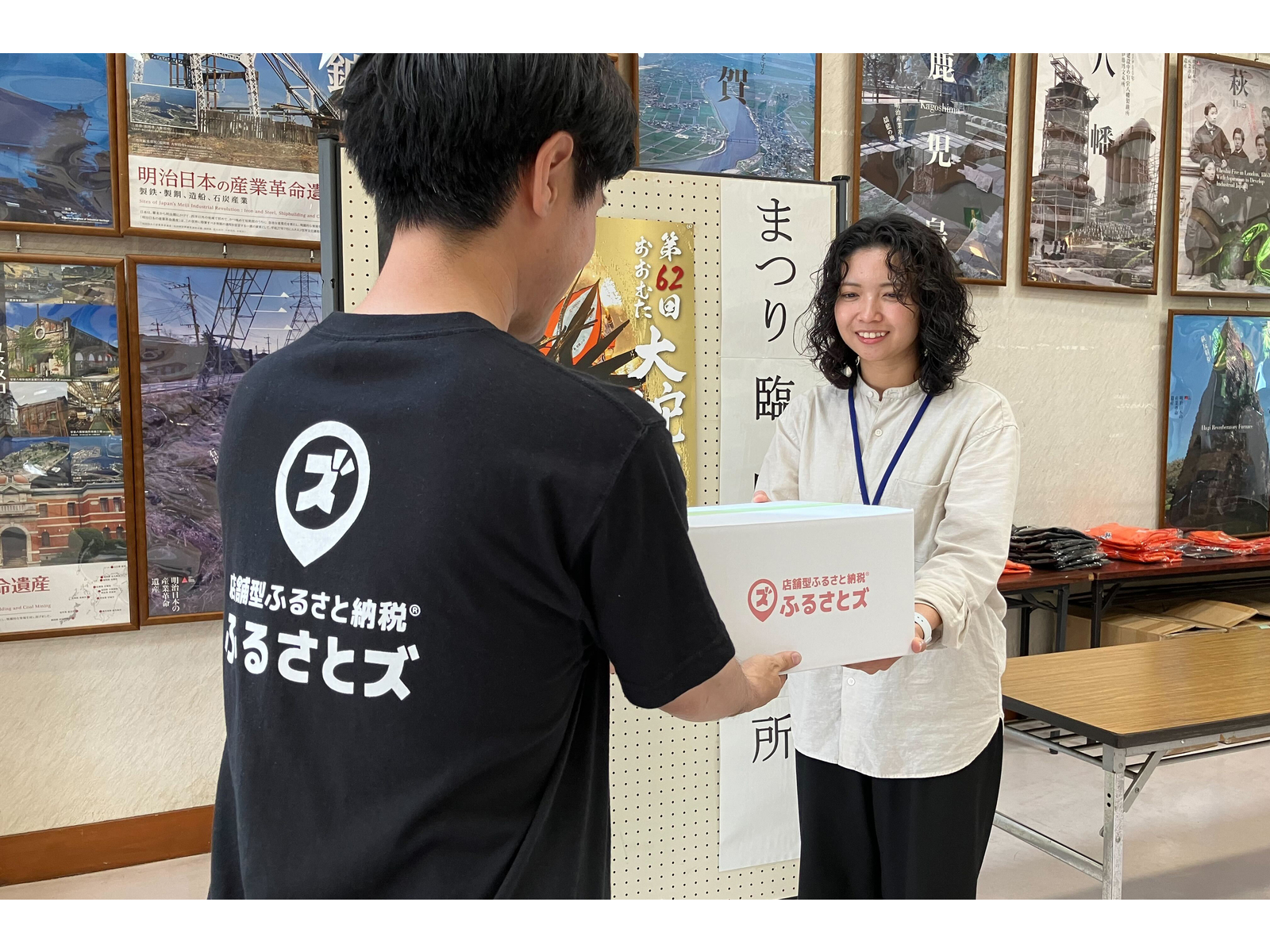 「ふるさとズ」のTシャツを着た男性が、「ふるさとズ」のロゴ入りの箱を女性に手渡すところ