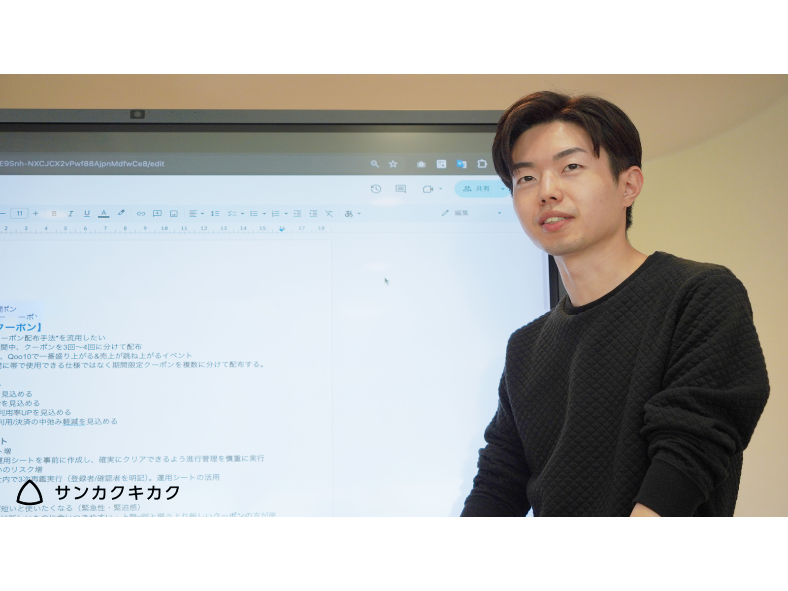 クーポン施策の企画書を映した大型モニターの横に立つ男性