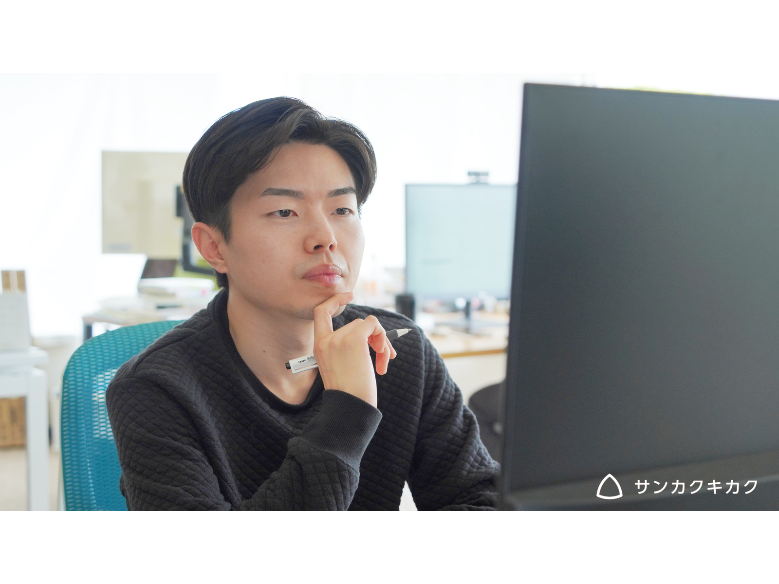 ペンを持ってあごに手を当て、モニターを見つめる男性