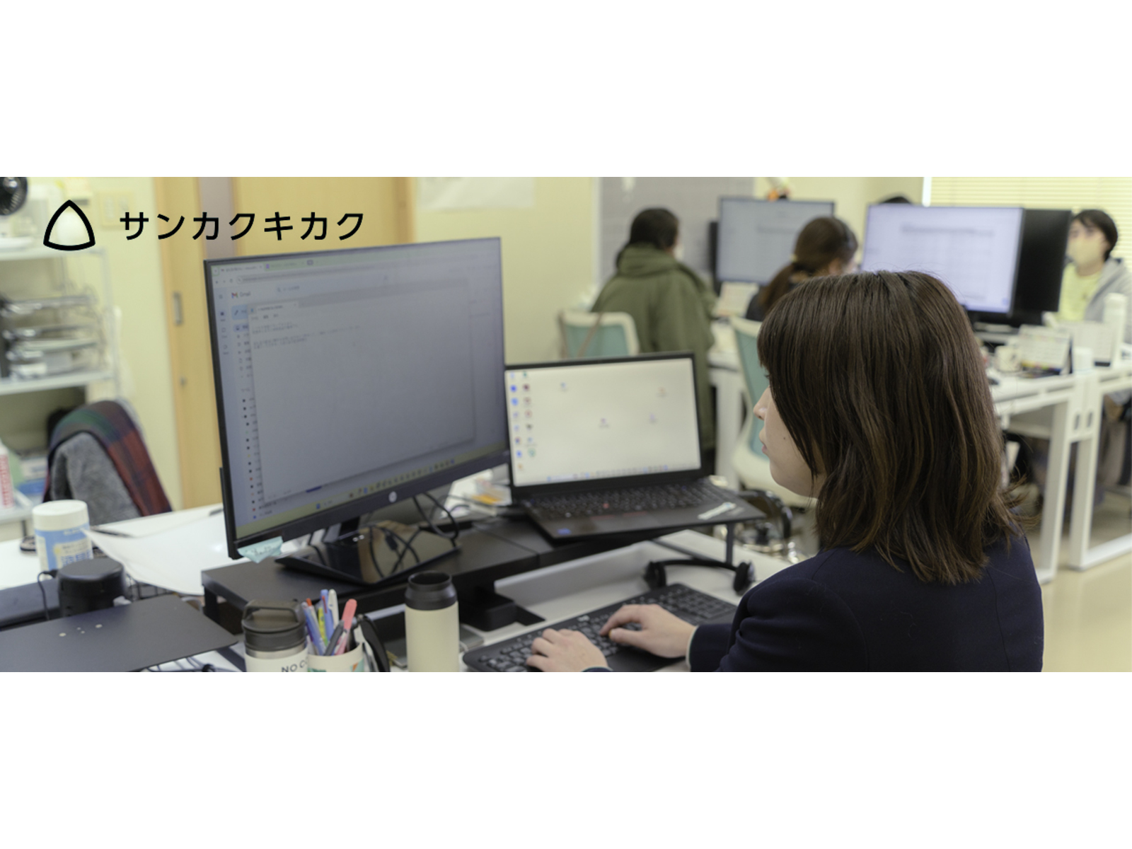 モニターとノートパソコンを並べた席でキーボードを打つ女性と、奥で作業する社員たち
