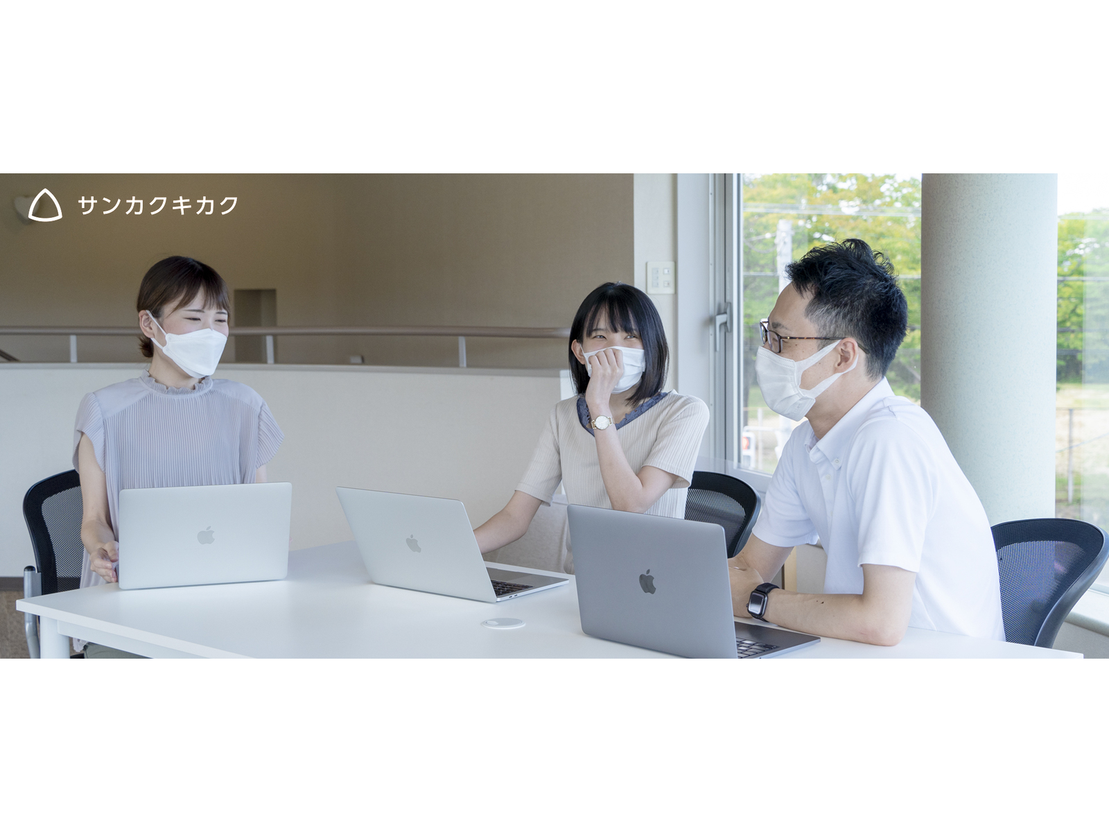 窓際の白い机でノートパソコンを並べ、和やかに話すマスク姿の社員たち