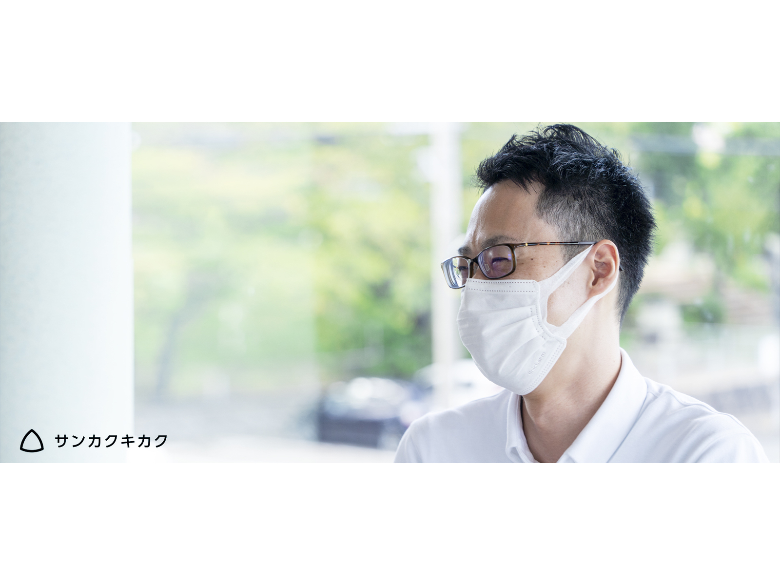 緑の見える窓辺で、横を向いて話を聞く眼鏡とマスク姿の男性