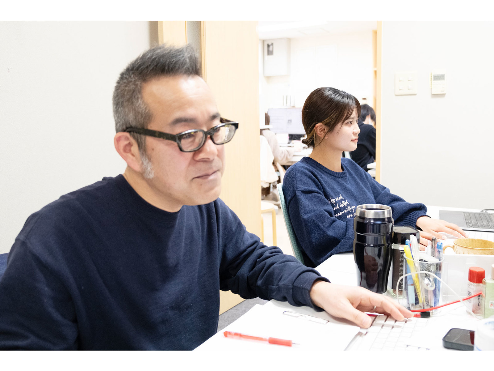 机に並んで座り、画面に向かって作業する紺のセーターの男性と女性