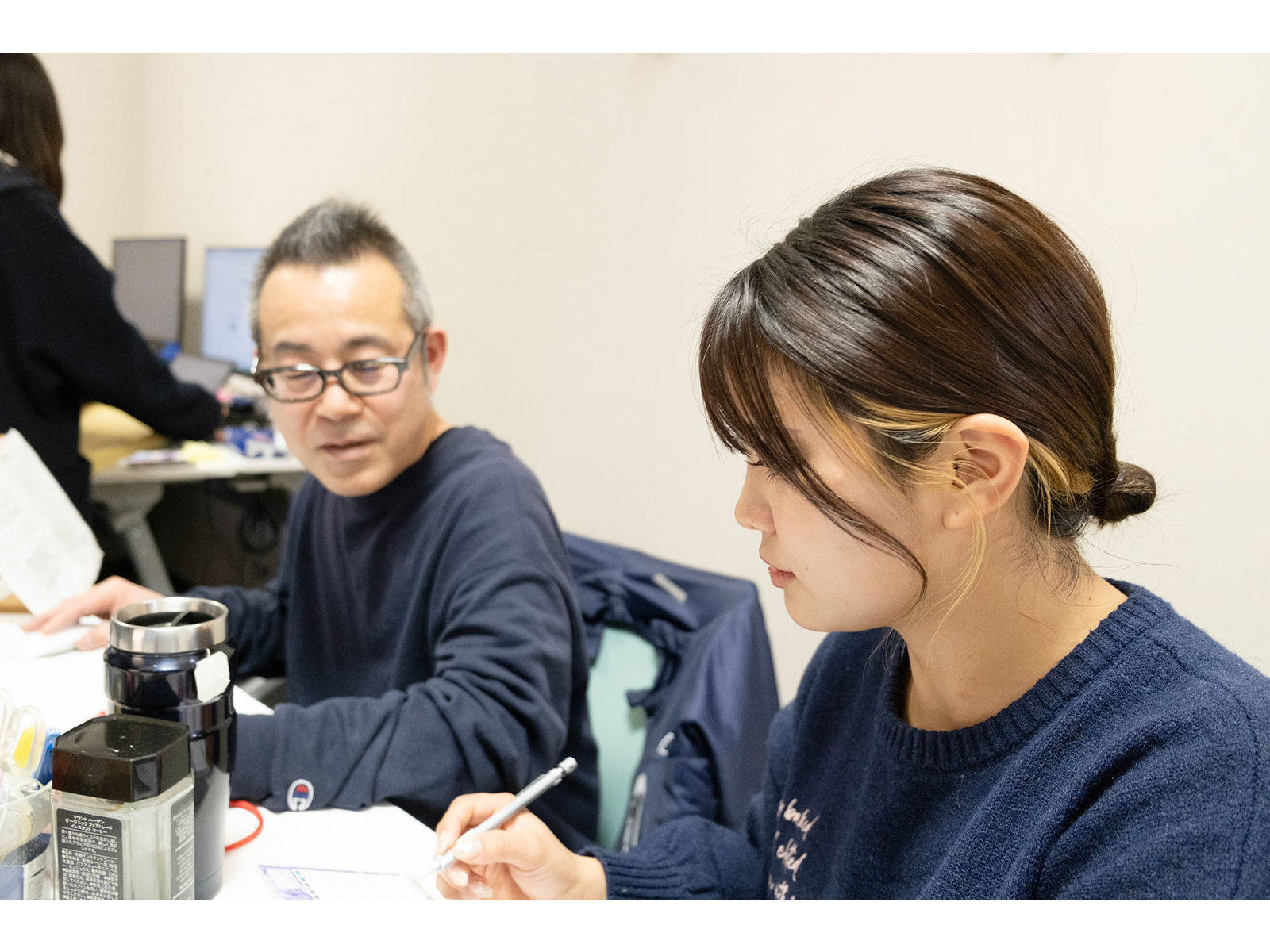 机で資料に書き込む女性と、その様子を横から見守る眼鏡の男性