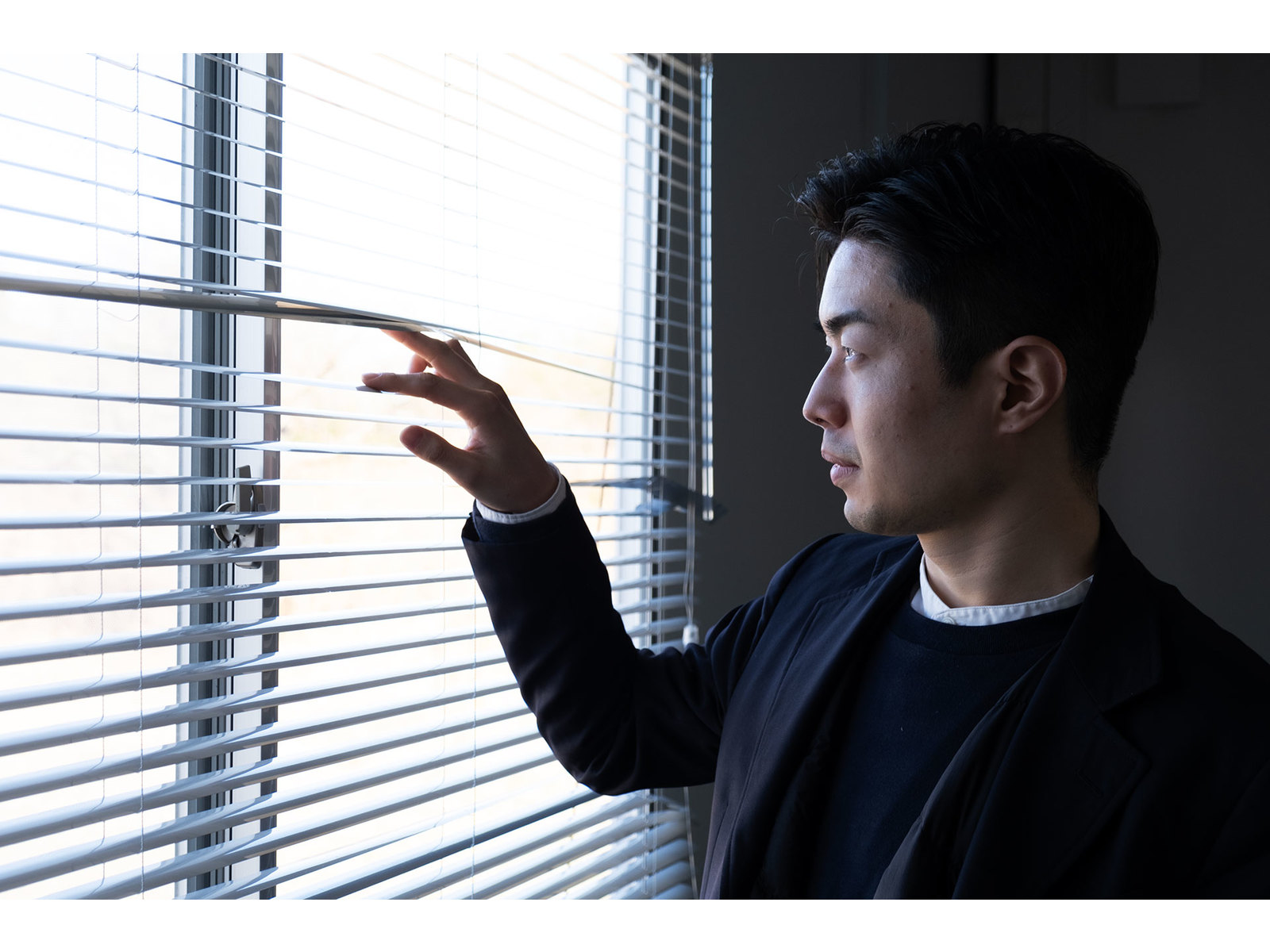 ブラインドを指で押し下げ、窓の外を見つめるジャケット姿の男性