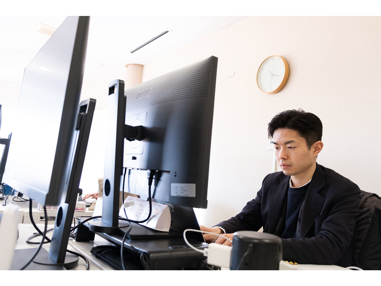 壁掛け時計のあるオフィスで、モニターに向かってキーボードを打つジャケット姿の男性