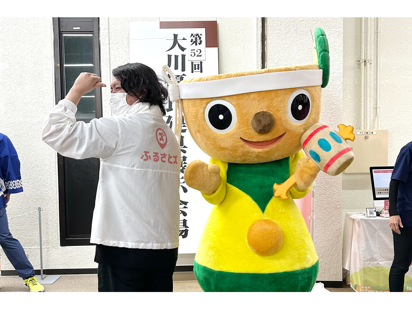 「第52回」と書かれた看板の前で、木づちを持つ着ぐるみのキャラクターと並んで力こぶのポーズをとる「ふるさとズ」の上着の人