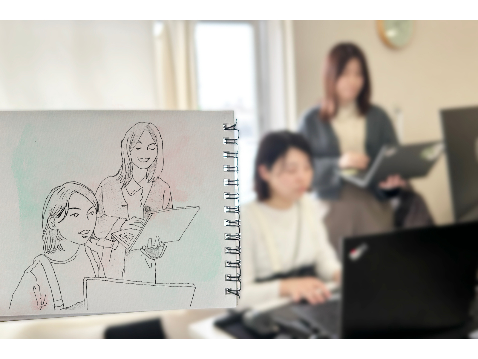 ノートパソコンで作業する女性たちを背景に、2人がパソコンを見ながら話す姿を描いたペン画スケッチ