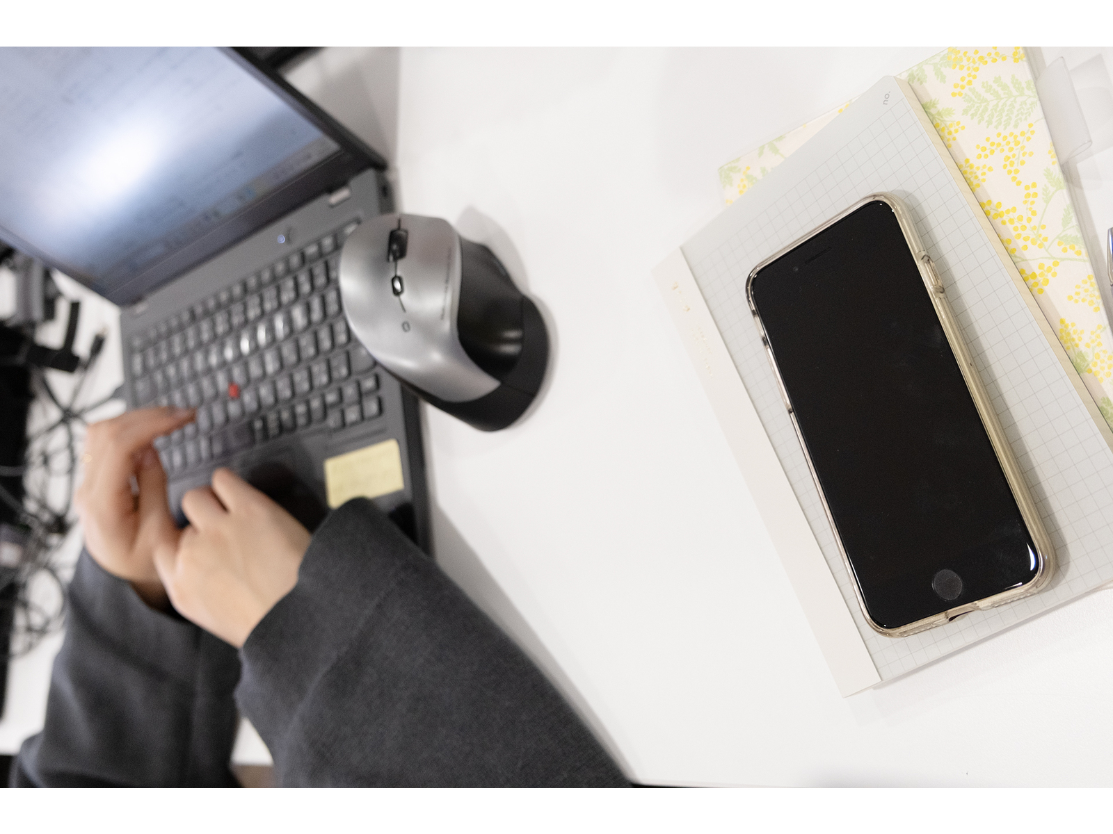 ノートパソコンのキーボードを打つ手と、机に置いたマウスやノートの上のスマートフォンを上から見た様子