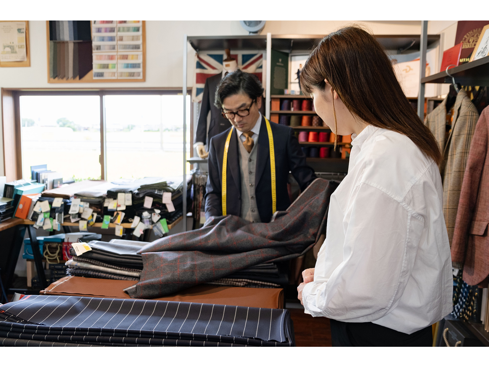生地が積まれたテーラーの店内で、メジャーを首にかけた店主の広げた生地を見る女性