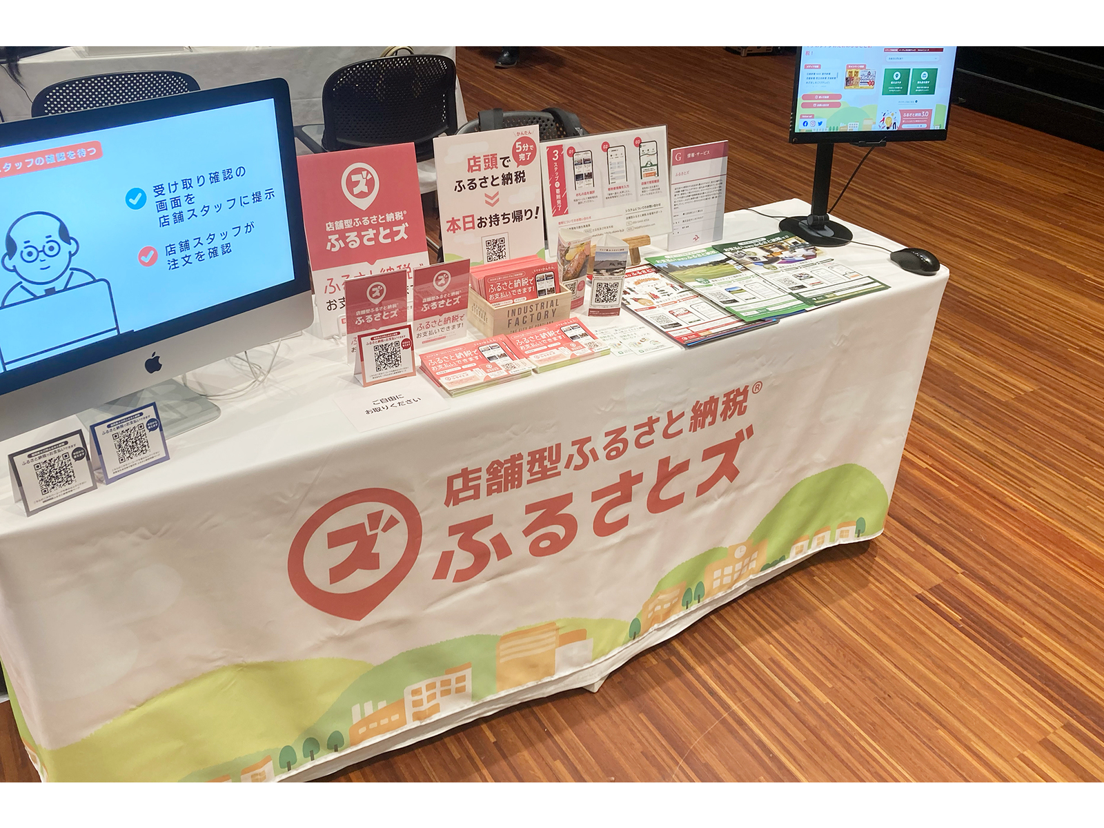 「店舗型ふるさと納税 ふるさとズ」のテーブルクロスを掛け、パンフレットやQRコード、説明画面のパソコンを並べた展示ブース