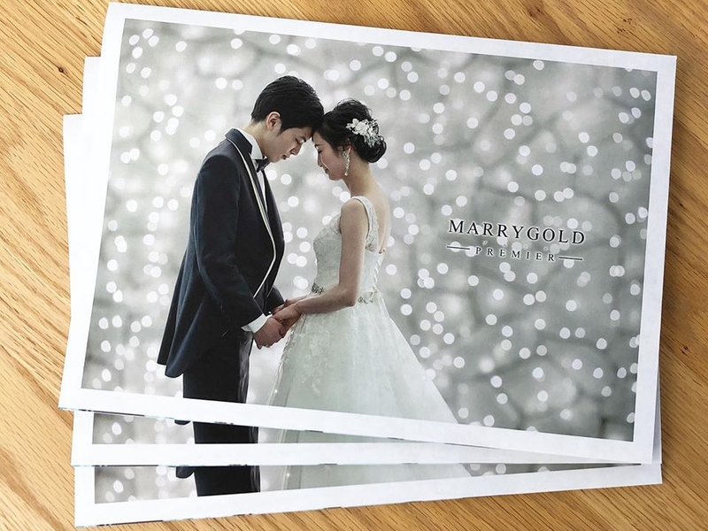 木の机に重ねたパンフレット。光の粒の背景で額を寄せ合う新郎新婦の写真に「MARRYGOLD PREMIER」の文字