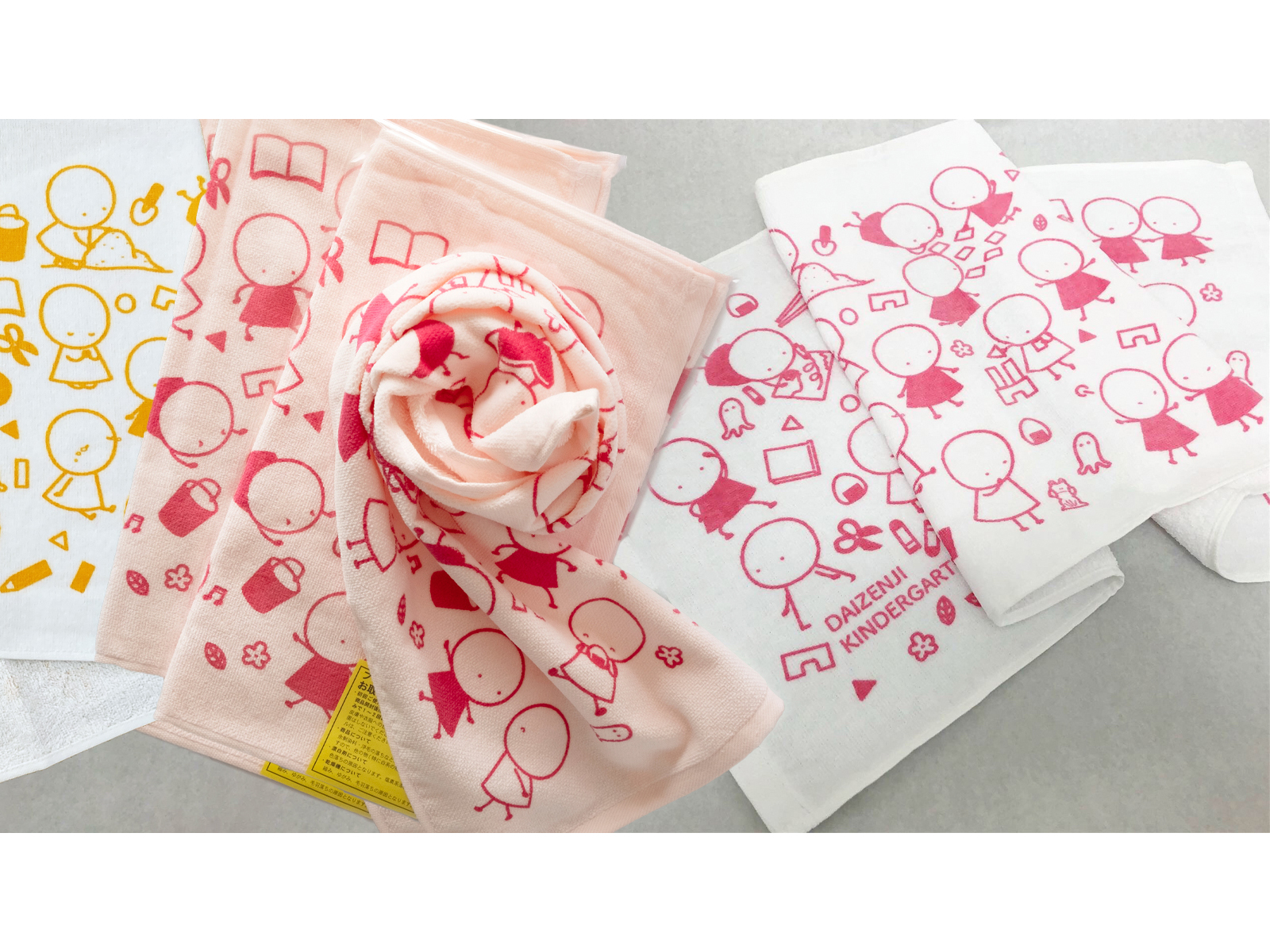 子どもや文房具の線画をプリントしたタオル数枚。黄色・ピンク・赤の柄があり、1枚に「DAIZENJI KINDERGARTEN」の文字