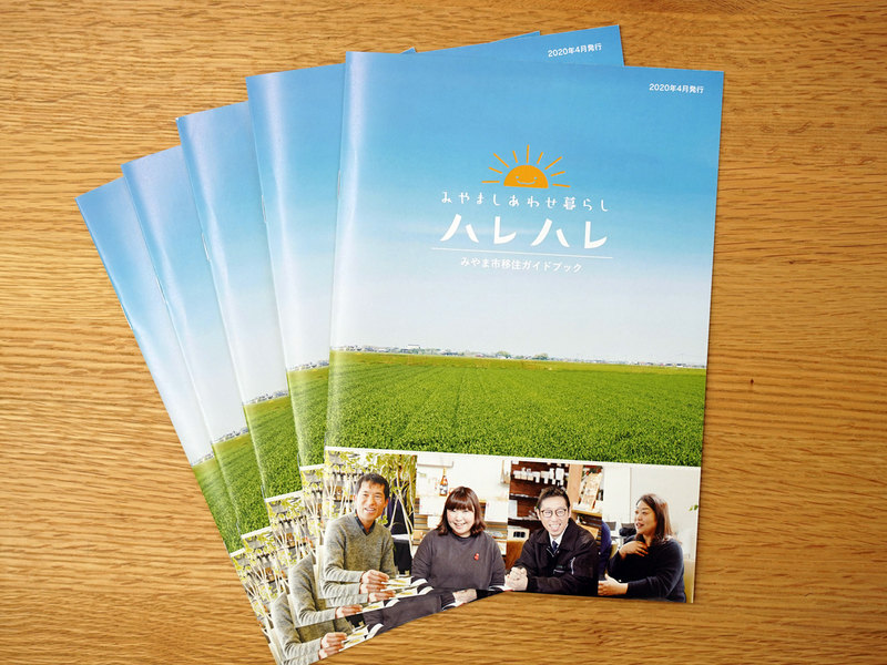 木の机に広げた移住ガイドブック5部。青空と緑の田畑の写真に「みやましあわせ暮らし ハレハレ」、下に笑顔の4人の写真