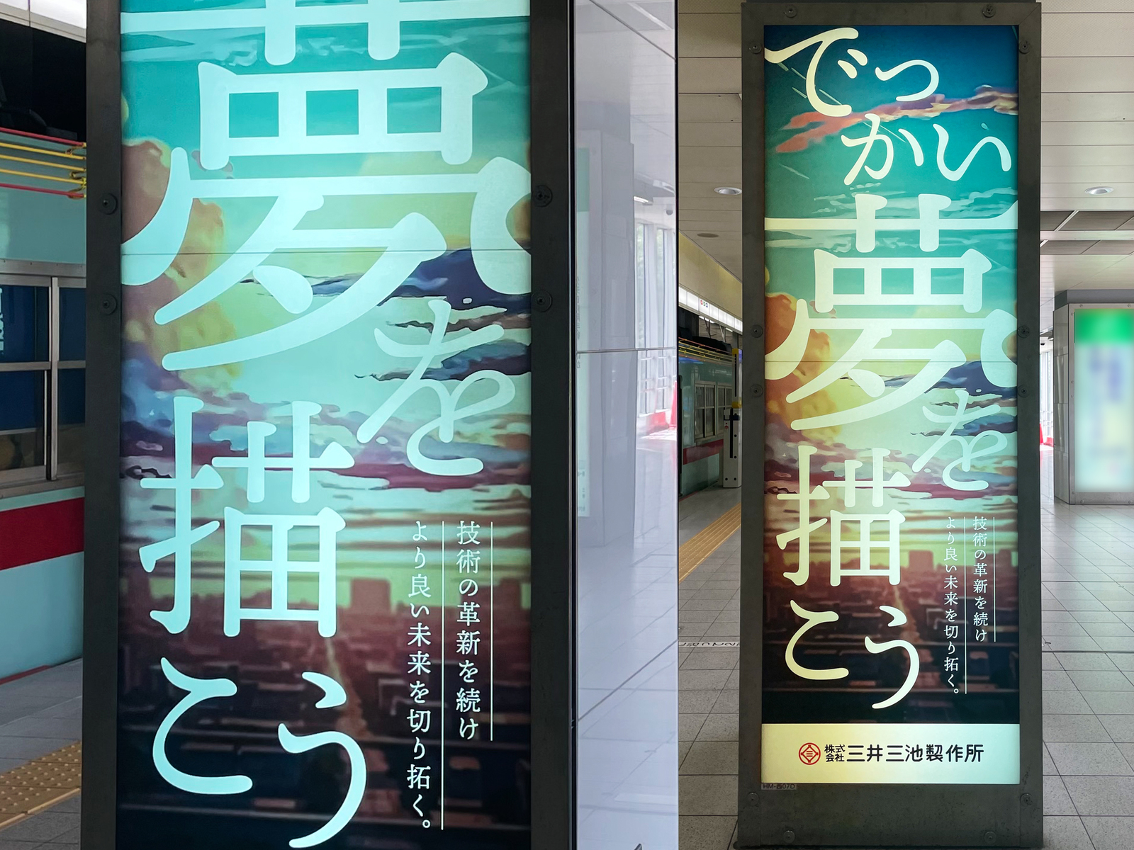 駅のホームの柱型サイネージ2台に映る縦長のグラフィック。夕焼けの街に「でっかい夢を描こう」「技術の革新を続けより良い未来を切り拓く。」の文字