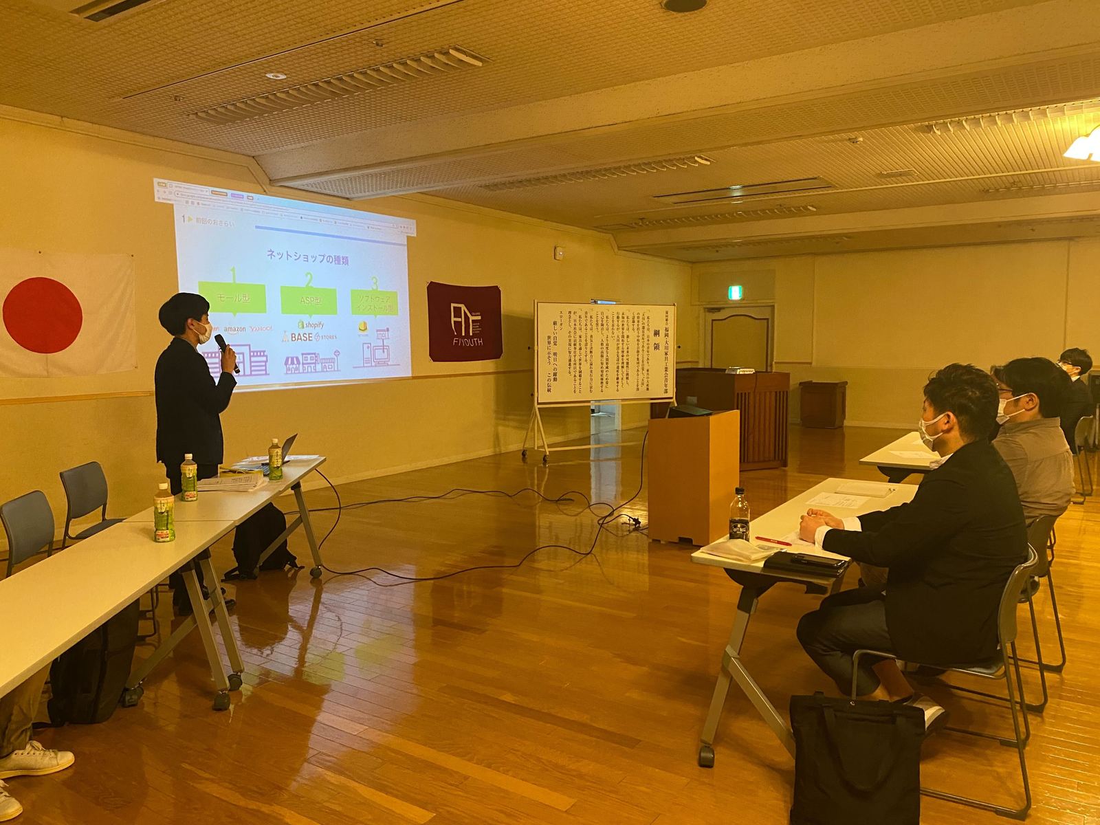 会議室のスクリーンに「ネットショップの種類」のスライドを映して話す講師と、机に座って聞く参加者たち