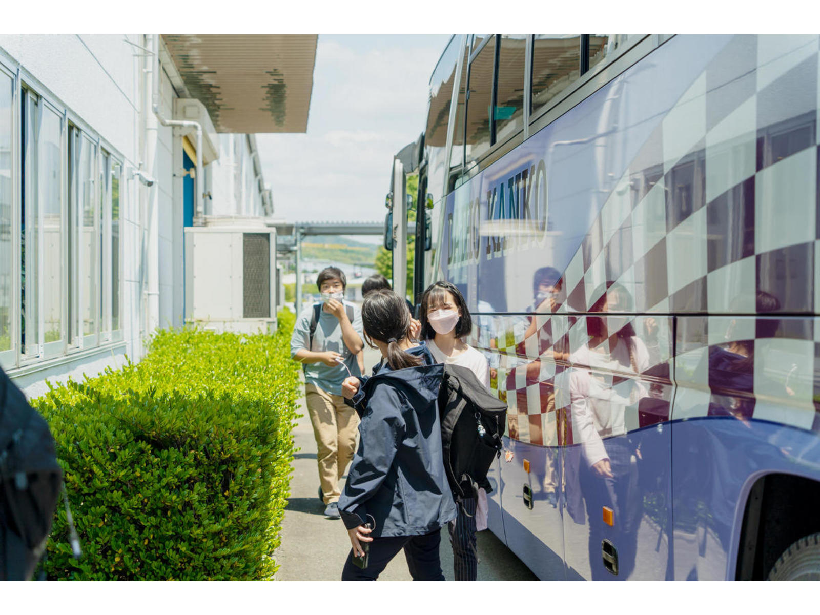 校舎の横に停まった大型バスの前を、リュックを背負って歩くマスク姿の学生たち